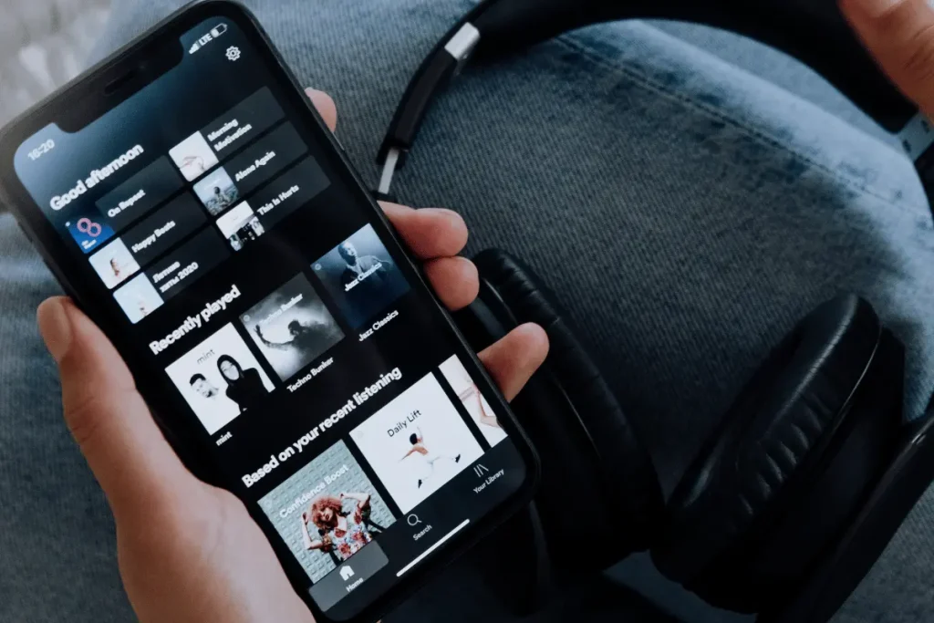 A hand holds a smartphone displaying a dark-mode music app’s playlists beside black over-ear headphones, suggesting relaxed music listening on the go.