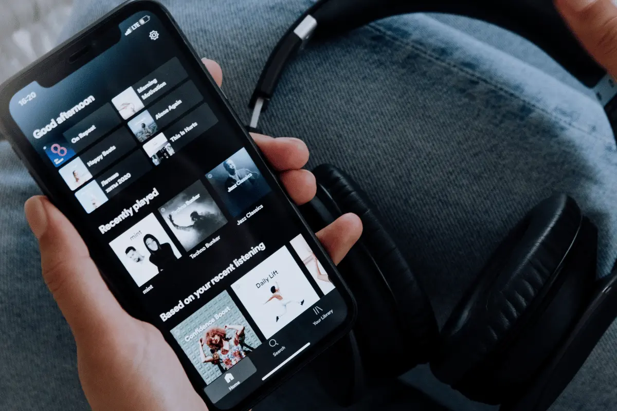 A hand holds a smartphone displaying a dark-mode music app’s playlists beside black over-ear headphones, suggesting relaxed music listening on the go.