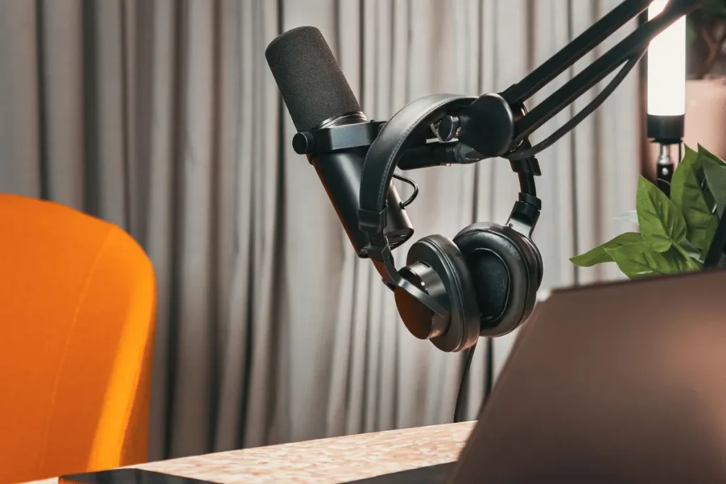 A studio microphone and headphones hang from a boom arm above a desk beside a laptop, with an orange chair and plant creating a calm podcast setup.