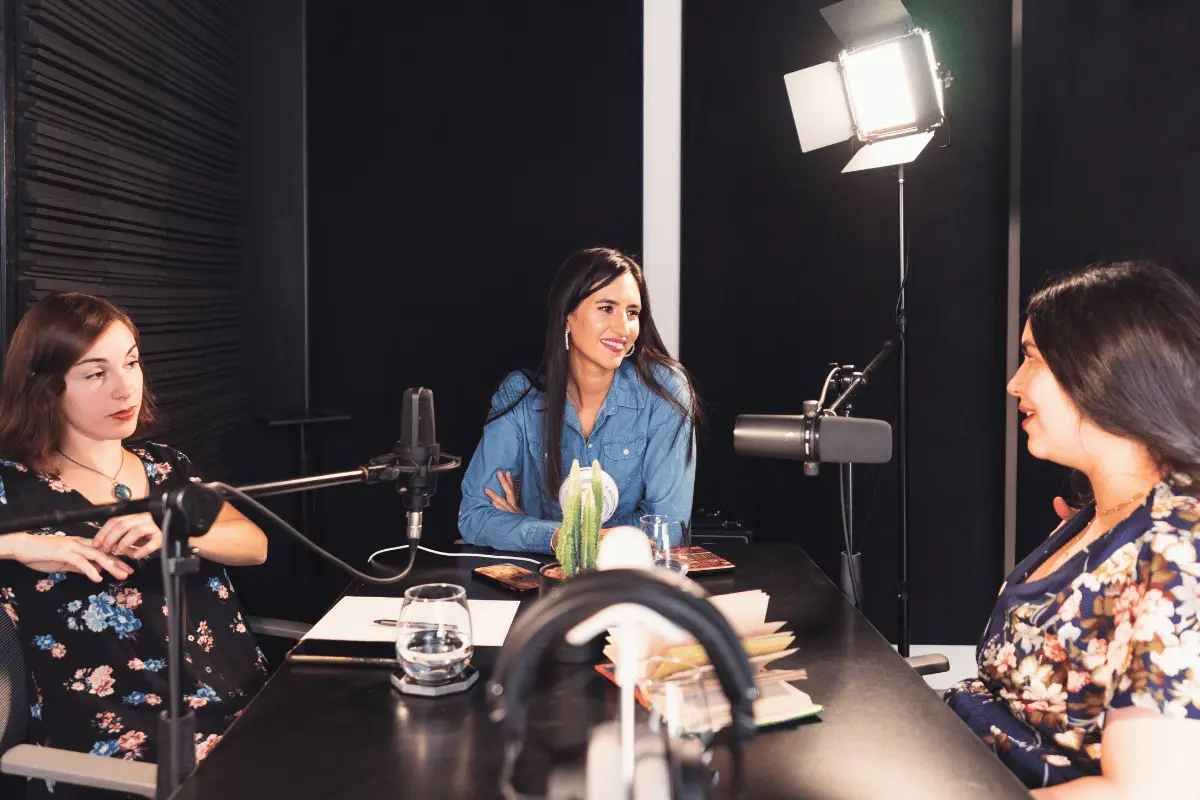 Three women in floral tops and a blue shirt chat around a studio table, with microphones and a bright light visible; the podcast setting feels warm and lively.