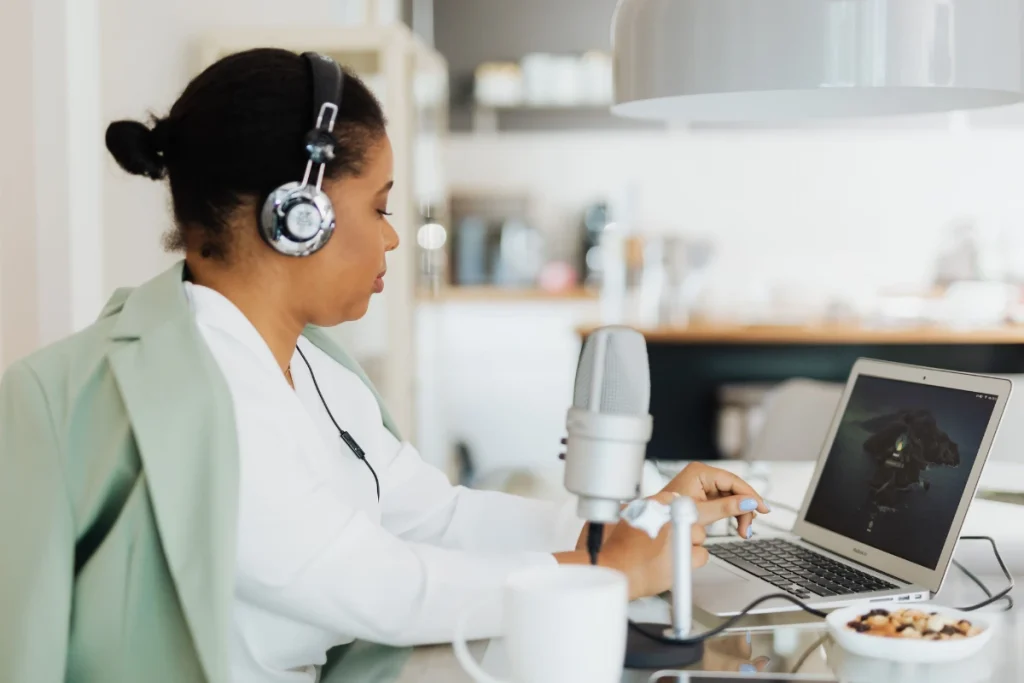 A woman wearing headphones works at a laptop beside a studio microphone in a bright kitchen, reaching toward its controls; calm, focused podcast recording.